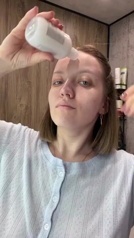 A young woman with short brown hair and blue eyes is shown applying a skincare product to her face. She is wearing a light blue, textured long-sleeved top and has light pink nail polish on her fingers. A gold ring is visible on her right hand, along with a gold chain necklace and rectangular earrings.

The woman holds a white plastic bottle, from which she dispenses a white liquid onto her forehead and then her cheeks. She uses her fingers to gently spread the product across her face. Her expression is focused and neutral.

Behind her, a wooden panel wall forms the backdrop, suggesting an indoor setting. To her right, a shelf is visible, holding several bottles and containers, indicating that the scene is likely taking place in a bathroom. Part of the text "catrix" is visible on the white bottle she is using.