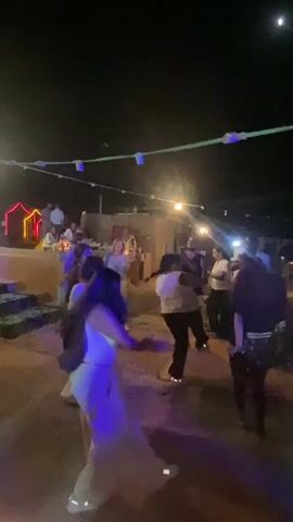 The video shows a group of people dancing outdoors at night. String lights hang above, casting a warm glow on the scene. Several individuals are captured mid-movement, their bodies blurred by the motion. In the foreground, a person in white pants and a dark jacket spins, their long dark hair flying. Other dancers in the background are also in motion, some with arms raised. The environment appears to be a somewhat rustic outdoor area, possibly a patio or courtyard, with structures visible in the background. The lighting suggests it is nighttime, with artificial lights providing illumination.