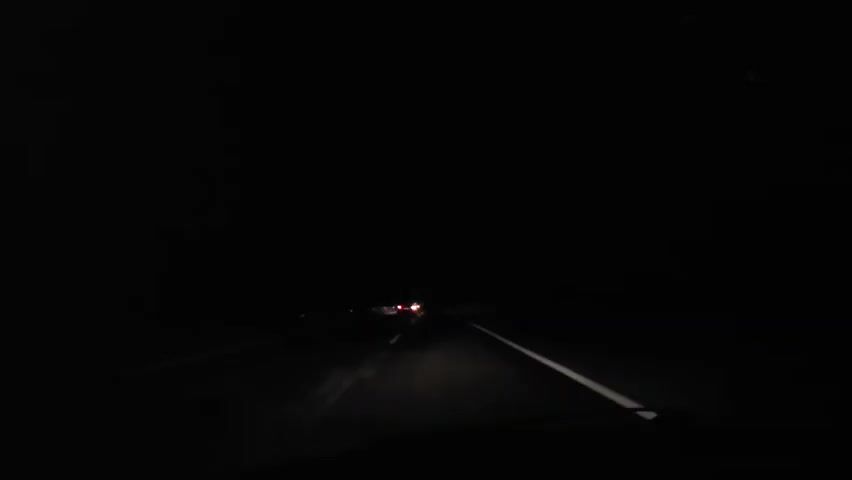 The video shows a first-person perspective from inside a car driving on a dark road at night. The car's headlights illuminate the road ahead, revealing a white solid line on the right and a white broken line on the left, indicating separate lanes of traffic. In the distance, several pairs of red taillights from other vehicles are visible, suggesting other cars are traveling ahead. The overall environment is completely dark, with no visible details of the surroundings beyond the road and distant taillights. The sky is black. There are no people visible. The action consists solely of the car moving forward on the road.