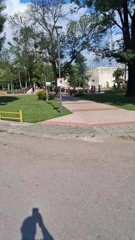 A daytime, eye-level, handheld, point-of-view shot pans left, starting from a public park with trees, a paved walkway, and a "CCTV" sign. The camera passes by a small patch of grass, a yellow metal fence, and a cracked asphalt path. A large, leafy tree dominates the mid-ground as the camera completes its leftward pan. The sky is bright blue with scattered clouds.