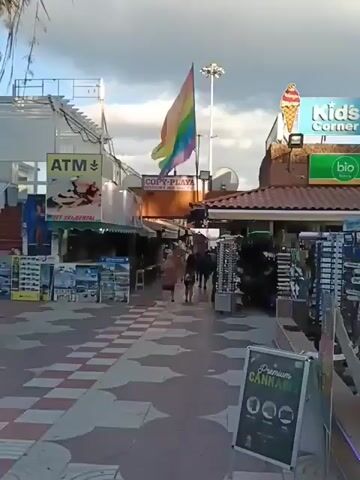 The video shows a street scene in a touristy area. There are shops on either side of a pedestrian walkway. One shop has an ATM sign and advertises jet ski rental. There's also a sign for "Copy-Playa" and a rainbow flag. Another shop sells "Premium Cannabis". People are walking along the walkway. On the other side of the walkway is a grass area with a trash can and a row of shops including an outlet store, a pharmacy, and a clothing store. A tall palm tree partially blocks the view. The sky is cloudy.
