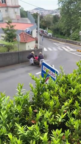 From an elevated vantage point behind green foliage, the video captures a street scene in a European town, possibly in Portugal given the Portuguese word "ATENÇÃO" (Attention) on a road sign. The street slopes downwards, with two people in white pants and dark shirts navigating a wicker toboggan across a crosswalk. A third person is seated in the toboggan. They are in motion, sliding down the hill.  On the left side of the street is a building with a red-tiled roof, a "P" parking sign, and a basketball hoop. A crowd of people is visible further up the street, suggesting onlookers or others participating in the toboggan activity. In the background, a cityscape unfolds with buildings and hills fading into the distance under an overcast sky. The focus of the video is on the toboggan's descent and the people involved.
