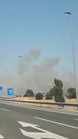 The video shows a view from a car traveling on a highway. On the left side of the road, there is a blue traffic sign with white arrows indicating three lanes. Beyond the highway barrier, there are trees and a large plume of dark smoke rising into the sky. The smoke appears to be originating from a location further in the distance. The sky is clear and blue, and it is daytime. The overall environment suggests an outdoor, possibly rural or semi-urban setting, with the smoke indicating a potential fire or other industrial event.