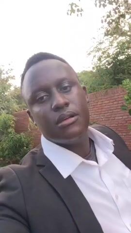 A medium shot shows a man wearing a white collared shirt and a black blazer standing outdoors. He is dark skinned and his hair is cut short. He is facing the camera and speaking. His right hand is slightly raised in front of him. Behind him there is a brick wall and some foliage. The video is recorded at a high angle and is slightly shaky.