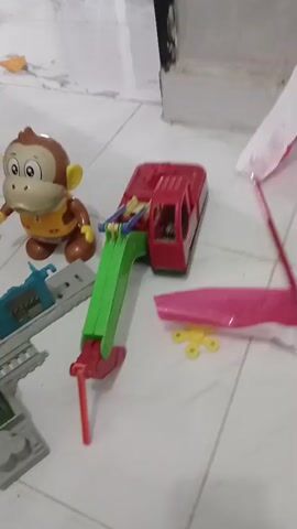 The video shows a collection of children's toys scattered on a tiled floor. In the foreground, a red toy excavator with a green arm is visible. To the left of the excavator, a brown toy monkey with a yellow vest is standing. Further to the left, a grey and blue toy train track piece is present. In the middle ground, a yellow fidget spinner lies next to a pink plastic sheet. Behind the excavator, two flags, one white and one red, are attached to a stick. In the background, the corner of a room is visible, with a dark door frame and white walls. As the video pans, more toys are revealed, including a green toy truck, a yellow toy excavator, and a black and red remote-controlled car with red wheels. The lighting is dim.