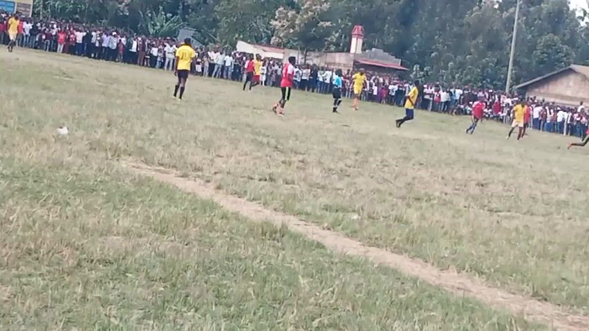 The video shows an outdoor event, likely a soccer match, on a grassy field. A crowd of people is gathered along the perimeter of the field, watching the game. Several individuals in athletic wear, some in yellow and others in red, are actively playing on the field. The background features trees, buildings, and a tent. The grass on the field appears somewhat dry and patchy, with a white line visible, possibly marking a boundary of the playing area. The overall impression is of a community gathering focused on a sporting event.