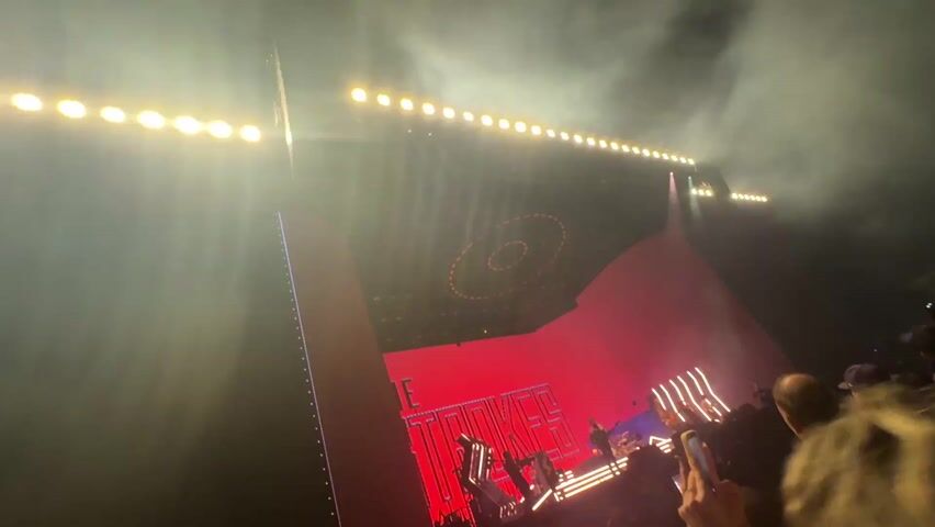 The video captures a rock concert in progress. Stage lighting is prominently displayed, with beams of light projecting into the audience.  Multiple large screens flank the center stage, showing images of people, potentially the band members.  Silhouettes of band members are visible on stage amidst the bright lights. The audience is packed closely together, their arms raised, many holding phones to record the performance. The overall atmosphere appears lively and energetic.