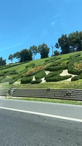 The video shows a view from a moving vehicle, likely a car, looking out the passenger side window and windshield. The time stamp indicates the video is approximately 4 seconds long. The scene is a sunny day with a clear blue sky. The vehicle is on a road, and to the right, there is a steep grassy hill. The hill is landscaped with bushes and trees. There are tiered sections on the hill, possibly for drainage or aesthetic purposes. Some of the landscaping on the hill appears to be in geometric shapes, possibly topiaries or shaped hedges. A modern street light with solar panels is visible on the hill. In the foreground, the side mirror of the vehicle is visible, reflecting the sky and landscape. The road itself is paved and appears to be a highway or a main road with a white line marking the lane. The overall impression is a well-maintained urban or suburban landscape viewed from a moving vehicle on a bright, clear day.