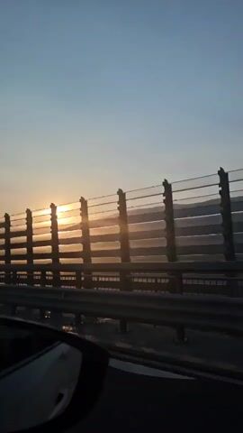 The video captures a view from inside a vehicle, likely a car, looking out towards a sunset. The sun is visible in the upper left quadrant of the frame, casting a warm, golden light. A highway sound barrier, consisting of vertical posts and horizontal panels, runs across the middle of the image, with the setting sun shining through its gaps. Beyond the barrier, distant hills or mountains are faintly visible through the hazy light. The foreground shows a portion of the vehicle's interior, specifically the side mirror and door panel, indicating movement. The sky is clear and a pale blue, transitioning to warmer hues near the horizon. The overall scene suggests a scenic drive during dusk.