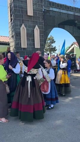 The video shows a group of people in traditional dress standing in an outdoor area. A large, archway structure made of dark material, possibly brick or stone, is in the background. The archway has a gothic architectural style with pointed openings. The sky is clear and blue, indicating daytime. Several individuals are wearing colorful, layered clothing typical of folk costumes, including long skirts, vests, and head coverings. One woman on the right holds a tall blue flag. People in the background are observing the group in traditional attire. The ground appears to be paved. The scene suggests a cultural event or festival.