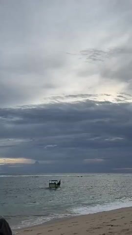 A lone boat drifts on the ocean near a sandy beach under a cloudy sky. The boat is small and appears to be anchored or adrift, with a canopy providing shade. The water is a calm, greenish-blue, with gentle waves lapping at the shore. The sand is light brown and appears wet near the water's edge. The sky is filled with a mix of dark and light gray clouds, suggesting either an overcast day or the prelude to a storm. The scene is serene and contemplative, with the vastness of the ocean and sky creating a sense of isolation.