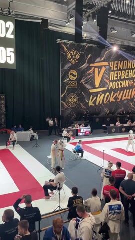 The video shows a martial arts competition taking place in a large indoor arena. Two competitors, dressed in white uniforms with colored belts, are engaged in a bout on a red and white checkered mat. An official in a black shirt monitors the fight. Surrounding the mat is a crowd of spectators, many wearing similar martial arts attire, along with coaches and officials. A large banner in the background displays the event name "Чемпионат Первенство России 2026 Кёкушин" (Championship of the Russian Federation 2026 Kyokushin). The arena is well-lit with multiple spotlights. Several other mats with similar competitions are visible in the background, and officials are seated at tables in the elevated area along the back of the arena.