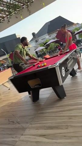 A man and a woman are playing pool on an outdoor pool table on a sunny day. The table is black with silver trim and a red felt top. A set of balls is racked on the table. The man wears a green polo shirt and black pants. He stands to the left of the table, holding a pool cue. The woman is wearing a pink bathing suit and white slide sandals. She stands to the right of the table, also holding a pool cue. In the background, there is a swimming pool, tables with red umbrellas, and a large house. The ground is tiled with a wood grain pattern. The camera is positioned at an angle to the table, and pans slightly to follow the woman as she moves around the table.
