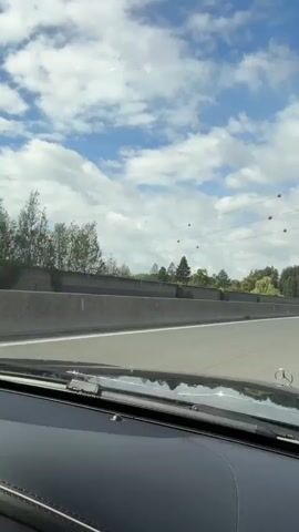 The video is a first-person perspective from inside a vehicle driving on a highway. The sky is blue with scattered clouds. Power lines with orange spheres are visible crossing the sky. A concrete barrier separates the highway lanes from the adjacent land, which features trees and what appears to be a sound barrier. Several other vehicles, including cars and a truck, are visible on the highway.