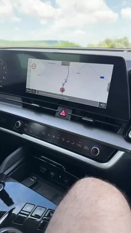The video begins with a close-up of a car's dashboard and navigation screen, showing a map with a route plotted. The perspective then shifts to a view of the road ahead through the windshield, with a bright, cloudy sky. The camera moves to show the interior of the car's door, then pans to the side-view mirror reflecting the passing scenery of a green, hilly landscape and the road. The scene appears to be a daylight drive on a highway.
