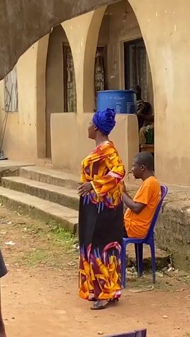 A woman in a vibrant orange and yellow patterned dress and a blue headscarf claps her hands while standing outdoors. She is facing away from the camera. Behind her, a man in an orange shirt sits on a blue chair. To the right of the man, another person is visible. Further in the background, a building with arched doorways is seen. Several people are gathered in the foreground and middle ground, some of whom are partially obscured by the camera's angle and foreground elements such as a tent or awning. The ground appears to be dirt. Blue chairs with text visible on their backs are placed in the foreground.
