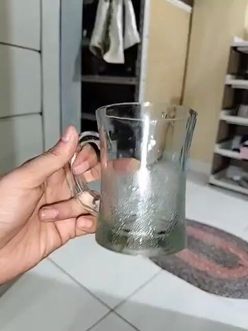 A person holds a clear glass mug with a textured exterior, filled with ice. The mug is condensation-covered. The person's hand, with visible fingernails, is gripping the handle. The background shows a tiled floor, a dark metal shelving unit with clothes hanging on it, and a woven rug. The lighting is dim.