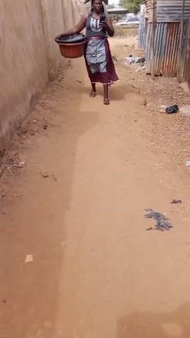 A woman walks down a dirt path in what appears to be an alley. She has a large bundle on her head and is carrying a large red tub. She is looking at a phone in her hand. The alley has walls on each side, one is a concrete wall and the other side is made of corrugated metal. There is trash strewn along the side of the metal wall. The camera follows the woman.
