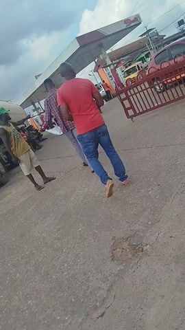 The video shows a man in a red t-shirt and blue jeans walking away from the camera on a paved surface. Behind him is a gas station with pumps and a canopy. There are other people and vehicles visible in the background, including a red car with orange stripes and a red metal barrier. The sky is cloudy.
