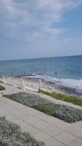 A panning video shows a view of a pebble beach with a calm, blue sea. Several people are visible in the water, some swimming and others wading near a pier. A white fence runs along the edge of a paved walkway lined with low-lying green plants. The sky is partly cloudy with a mix of blue sky and gray clouds. In the distance, a white building with a red roof sits atop a cliff overlooking the sea. The video pans from right to left, revealing more of the coastline and the buildings along it.