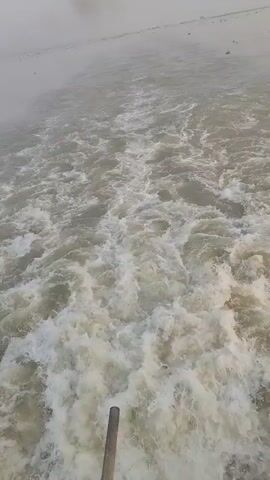 The video shows a view of water with many waves and white foam. The water is a murky grey color, and a small boat can be seen in the distance. The sky is overcast and grey. The camera is positioned above the water, looking back at the waves. It is a shot from a moving vessel.