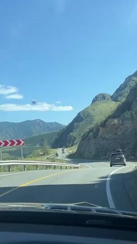The video shows a car's perspective on a winding road through a mountainous landscape. The road has a yellow dividing line and a white edge line. Several cars are visible on the road ahead, following the same route. The landscape consists of green hills and mountains under a clear blue sky with scattered white clouds. The camera is looking forward through the windshield.