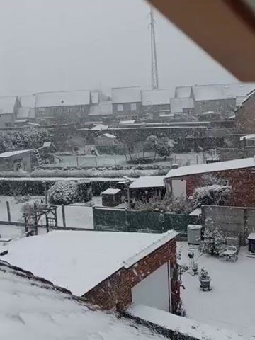 The video shows a snowy neighborhood from an elevated perspective, possibly a window or balcony. Snow is falling heavily, covering roofs, yards, and streets. Houses are visible in the background, with snow-covered roofs and facades.  A backyard is visible with snowy furniture and shrubbery. A street is also visible covered in snow with parked cars.