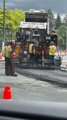 The video shows an asphalt paving operation on a road. A large paving machine, likely an asphalt paver, is in the center of the frame, laying down a fresh layer of asphalt. A dump truck is positioned behind the paver, its bed tilted up, feeding asphalt into the paver. Several construction workers are visible. Three workers stand to the left of the paver, one holding a long-handled tool. Two workers are on the paver itself, operating the machinery. One worker is positioned on the right side of the paver. They are all wearing safety vests and hard hats. The environment appears to be a suburban or semi-rural setting, with trees and power lines visible in the background. The sky is overcast. Orange traffic cones are placed on the road, indicating a work zone. The asphalt is steaming as it is being laid.