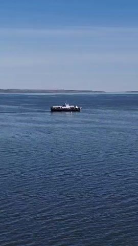 The scene is a long shot of a large body of water under a blue sky with faint, wispy clouds.  In the middle distance, a ferry boat sits motionless on the water. The ferry has a flat deck, with a white cabin structure at the front. The water is rippled.  The far shore is visible as a dark line on the horizon. An off-screen voice speaks.