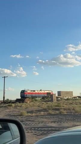 The video shows a train moving from left to right across a dry, grassy landscape under a bright blue sky with scattered white clouds. The train is composed of a red, white, and blue locomotive followed by several flatbed train cars carrying large, rectangular containers. One of these containers is labeled "CDRS". To the right of the train tracks, there is a small, weathered wooden structure that resembles an outhouse. In the foreground, a portion of a car's side mirror is visible, indicating the video is likely being filmed from inside a vehicle. A utility pole with wires is visible on the left side of the frame, and various pieces of debris, possibly discarded wood or metal, are scattered near the tracks. The overall environment appears to be rural and arid.