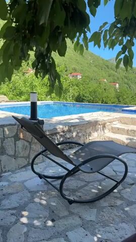 The video shows an outdoor scene with a rocking chair in the foreground. The chair is made of dark grey fabric and has a black metal frame. Behind the chair is a swimming pool with blue water, surrounded by a stone wall. Further in the background, there are lush green trees and a mountainous landscape. The sky is clear and blue. The overall impression is one of a peaceful and relaxing environment.