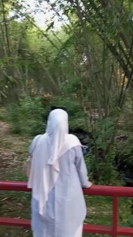 A person wearing a light-colored hijab and a matching outfit stands on a red-railed bridge, looking at a bamboo forest. A small stream flows beneath the bridge, surrounded by lush greenery. The camera pans to the right, revealing more of the dense bamboo forest and the path that winds through it. The sun filters through the leaves, creating a dappled light effect.