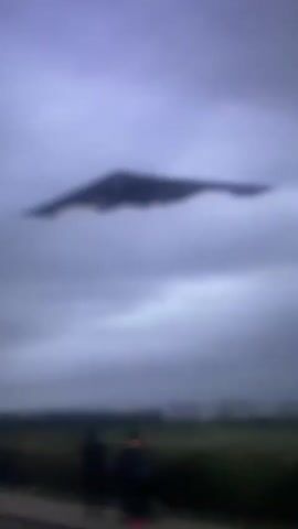 A black, stealth bomber flies through a cloudy sky over a dark, horizontal landscape. The bomber is dark and appears to be a B-2 Spirit, a flying wing design. The underside of the bomber is visible, and it appears to have landing gear extended. The sky is overcast and appears to be during the daytime, though the light is diffused. The landscape below is dark and appears to be a mix of fields and trees, with no clear details visible due to the lighting and distance. The video is shaky and the focus is soft, indicating it was likely filmed handheld at a distance.