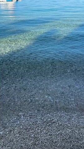 A short video captures a close-up view of a tranquil beach. Crystal-clear turquoise water gently laps at a pebble-strewn shore. The sunlight casts dappled shadows on the seabed, which is visible through the transparent water, revealing a mosaic of smooth, light-colored stones. The water is calm, with only a hint of ripples and small waves breaking near the edge. In the upper left corner of the frame, a hint of a white structure, possibly a building or boat, is visible against the blue sky. The overall impression is one of peace and natural beauty.