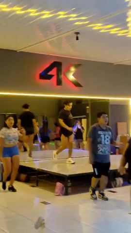 This video shows a group of people, appearing to be adults and young adults, participating in what looks like an energetic dance or aerobics class. They are moving their arms and legs in synchronized movements, some on a raised platform or stage, and others on the floor around it. The setting is an indoor studio or gym with artificial lighting, including decorative yellow patterned lights on the ceiling and a prominent, bright red neon sign on the back wall that reads "4K." A large mirror behind the stage reflects the participants and the room. The individuals are dressed in casual workout attire, such as shorts and t-shirts. They appear engaged and energetic during the activity. The location is an indoor facility, possibly a fitness center or dance studio in Tacna, Peru, as indicated by the context.