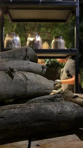 The video captures an evening scene, likely outdoors or in an open-air restaurant setting in Nir Eliyahu, Israel. In the immediate foreground, a stack of dark, weathered logs, possibly charcoal, rests on a wooden counter. Behind this, a chef, wearing a black t-shirt, a striped apron, black gloves, and a white kippah, is actively working. He is holding a white plate and appears to be preparing or serving food. A tall glass containing a dark beverage with ice is also on the counter near him. In the background, shelves hold various jars, and an active grill is visible with food cooking. Other individuals are present in the background, some appearing to be formally dressed, including one wearing a top hat. Trees are visible beyond the illuminated area. The ambient sounds include music and people talking, indicating a social gathering or event.