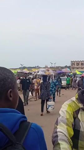 The video shows a bustling outdoor market in what appears to be an urban area. The sky is overcast. Many people are walking through the market area. Some individuals carry goods on their heads, while others carry bags or containers. Market stalls with colorful umbrellas and canopies are visible, suggesting a variety of goods being sold. Motorcycles move through the crowd. Buildings are visible in the background, including one with a minaret. 
The camera moves around, focusing on individuals and the overall market scene. The focus is primarily on the ground-level activity and the crowd of people.