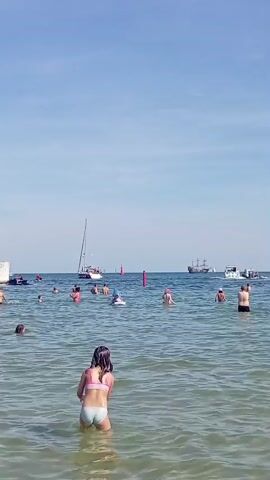 This video captures a lively summer scene at a beach in Gdynia, Poland. The primary subjects are numerous people of various ages enjoying the water on a clear, sunny day. Children and adults are seen wading, swimming, and playing in the gentle waves of the Baltic Sea.

In the foreground, a young girl in a light-colored bikini stands facing away from the camera, looking out at the sea. Another girl in a blue and white patterned swimsuit appears closer to the shore, splashing water. The water itself is a clear, greenish-blue, with sunlight glinting off its surface.

Further out in the water, a diverse group of individuals are scattered, some floating or swimming, others simply standing and conversing. In the background, several boats are visible. A sailboat with a white sail is prominent, as is a larger ship, possibly a tourist boat or a historical vessel, with multiple decks and masts. Smaller motorboats are also present, navigating the waters.

The setting is a coastal area, suggested by the presence of a pier or jetty on the left side of the frame, marked by a green navigational buoy and a white structure. Red buoys are also dotted in the sea. The sky is a bright, cloudless blue, indicating fair weather. The overall atmosphere conveyed is one of joy, relaxation, and summer fun. The time of day appears to be midday or early afternoon, given the bright sunlight and the shadows. No visible text is present in the video.