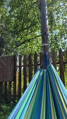 A colorful hammock hangs from a wooden pole in a yard. The hammock is striped with blue, green, and yellow. Behind the hammock is a wooden fence and green trees. The sun is shining.