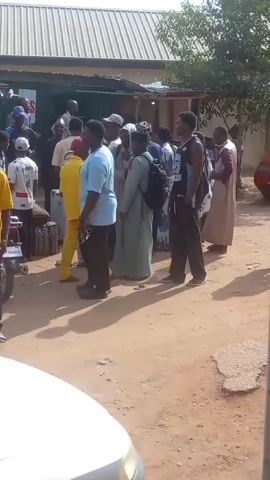 The scene depicts a crowded area with many people gathered in front of a building with a corrugated metal roof, likely a store or shop. The people are dressed in a variety of clothing, including casual wear, traditional attire, and uniforms. Some are carrying bags or packages. The environment appears to be outdoors, with a dirt or gravel ground and some trees in the background. There is also a white car visible in the foreground. The atmosphere seems busy and possibly bustling, suggesting a marketplace or a place where people are gathering for a specific purpose.