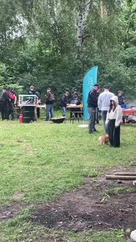 The video shows an outdoor gathering in a wooded area. Several people are present, some standing around tables, others near grills or food preparation areas. One person walks across the frame from right to left. A dog is also visible in the foreground. A teal flag with white lettering stands near some of the people. The ground is covered in grass and some dirt patches with scattered wood. Trees and foliage form a backdrop.