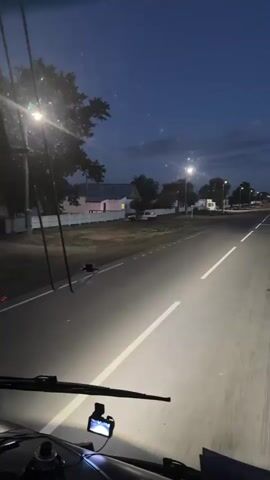 This media file captures a first-person perspective of a journey through Baiqadam, Kazakhstan, at dusk. The scene is a paved road illuminated by the headlights of the vehicle, suggesting travel is underway. The sky is a deep blue, transitioning from twilight, with faint clouds visible. Streetlights are spaced along the roadside, casting pools of light.

On either side of the road, there are glimpses of a rural or suburban setting. Houses with white fences and modest buildings are visible on the left, interspersed with trees. On the right, more buildings and open grassy areas can be seen. A speed limit sign displaying "20" is present on the right side of the road.

The overall atmosphere is one of quiet travel as day transitions into night. The lighting suggests that it is either early morning or evening. There are no people explicitly visible in the frame, but the presence of a vehicle and the organized nature of the road and buildings indicate human presence and activity. The media appears to be a video clip, given the timestamped frames. Some text is visible on the dashboard of the vehicle in the foreground, but it is largely illegible.