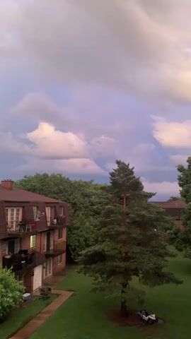 This is a still image of an exterior view of an apartment building. The building is made of brick and has multiple floors with balconies. There are several trees in the foreground and background. The sky is cloudy and appears to be at sunset or sunrise, with hues of pink, purple, and yellow. The ground is covered in grass. There is a walkway leading to the building. A white utility vehicle is parked on the grass near a tree.