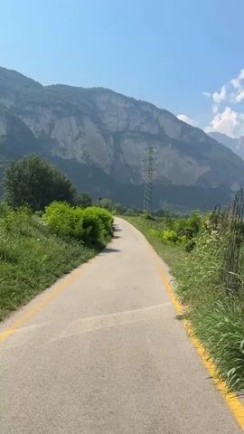 The video is a first-person perspective of someone cycling on a paved road with yellow lines on the side. The road curves to the left. On the left side of the road is a lush green bush. Further back, there is a large mountain range with sheer cliffs and sparse vegetation. A tall, lattice metal tower with power lines is visible on the mountainside. On the right side of the road, there is tall green grass and more distant mountains. The sky is clear blue with a few wispy white clouds. The sun is shining, creating shadows on the road. The overall environment appears to be a rural, mountainous landscape on a sunny day.