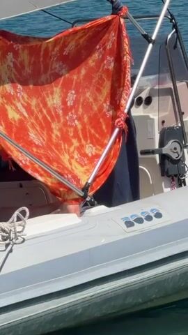 A vibrant, orange and yellow tie-dye sarong with white floral and turtle motifs is draped over a metal frame on a boat. The sarong is attached to the frame with red cord. The boat appears to be a dinghy or tender, with gray inflatable tubes. A white rope is visible on the left side of the boat. In the background, the blue water of a bay or ocean is visible. A person's legs are briefly visible behind the sarong, suggesting they are sitting or lounging on the boat. The sun is shining, creating bright light and shadows.