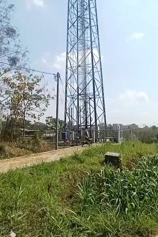The video shows a tall, lattice-style telecommunications tower in an outdoor setting. The tower is situated in a grassy area with some trees and foliage visible. A dirt path runs in front of the tower, and some electrical equipment is visible at its base. There are a few clouds in the sky, and the sun appears to be shining. The camera is positioned at ground level, looking up at the tower. The video is likely a short clip, possibly a pan or a stationary shot. No people are clearly visible in the video, although there might be a figure in the distance near the base of the tower in one frame.