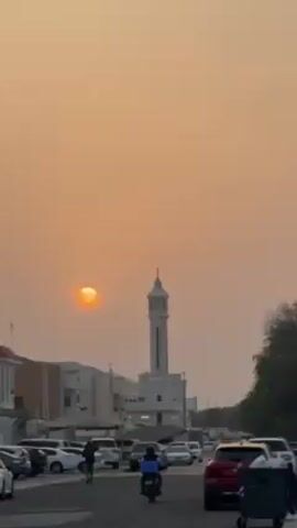 The video shows a view of a street at sunset. The sky is a hazy orange color, and the sun appears as a bright orange disk in the sky.  A white mosque with a tall minaret stands in the distance.  The street is lined with parked cars and buildings. Several people are walking on the street, and a scooter is driving.