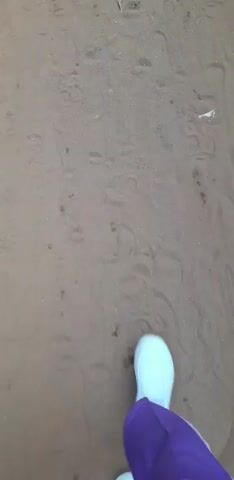 The video shows a first-person perspective of someone walking on a dirt ground. The ground is covered in footprints, and there are a few small sticks and stones scattered around. The person is wearing white sneakers and purple pants. The camera is focused on the ground in front of the person as they walk.