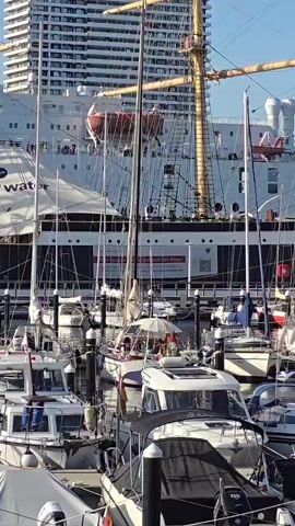 The video shows a harbor scene with several boats docked. In the background, a large white ship with a prominent mast and rigging is visible, along with a tall modern building. In the foreground, smaller motorboats and sailboats are moored. The lighting suggests it is daytime.