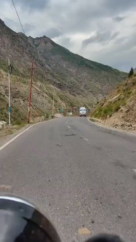 The video shows a highway winding through a mountainous landscape. The road is paved and appears to be in good condition. On the left side of the road, there is a steep, rocky mountainside with some sparse vegetation. There are also power lines and utility poles. On the right side, the mountainside is also steep and covered with vegetation. In the distance, a white truck with colorful decorations on its cab is traveling on the highway. The sky is cloudy, suggesting overcast or impending weather. The perspective suggests the video is filmed from a moving vehicle, likely a motorcycle given the visible portion of the vehicle's front.